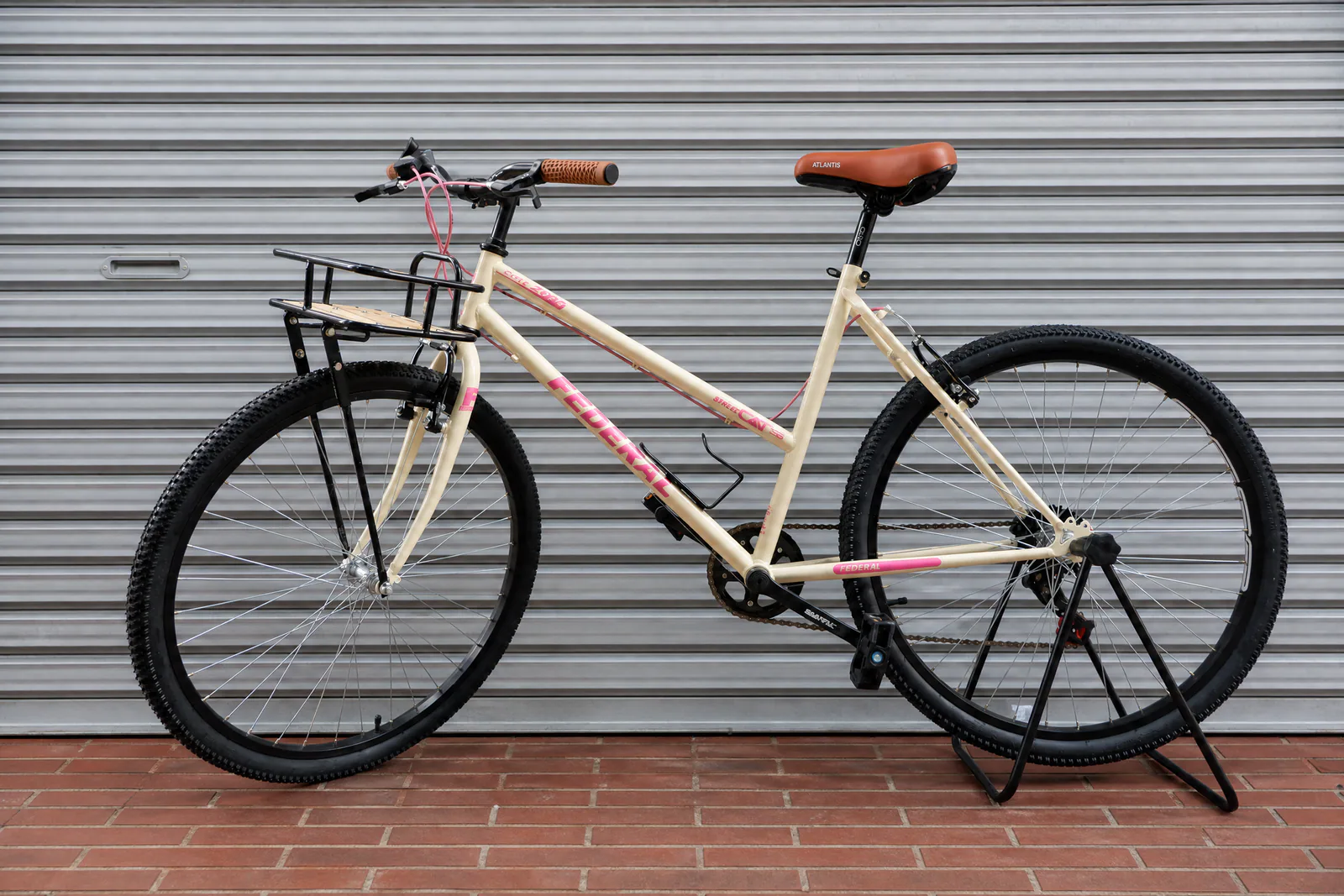 Sepeda Federal Lady warna cream dengan aksen pink, sadel dan grip cokelat, serta rak depan kayu, dipotret di depan rolling door abu-abu.